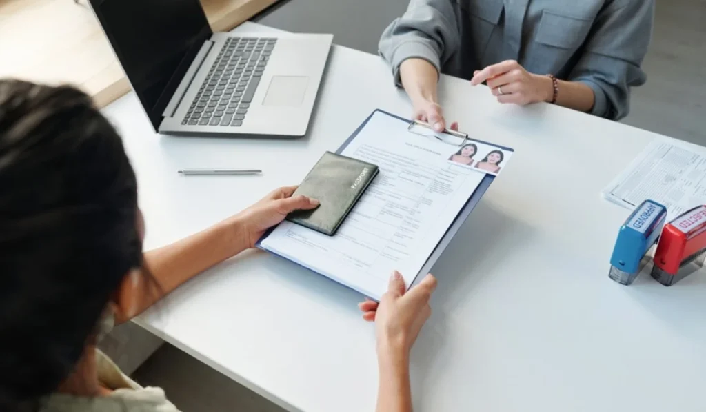 a women visa consultant passing the passport and some documents to the men and there is a laptop on the table.