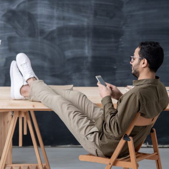 young-employee-in-casualwear-keeping-legs-on-table-while-scrolling-in-smartphone-1080x720