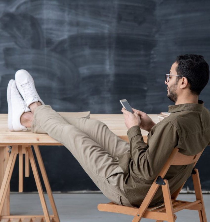 young-employee-in-casualwear-keeping-legs-on-table-while-scrolling-in-smartphone-1080x720